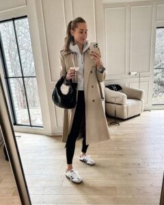 Kvinna i beige trenchcoat, svarta tights och sneakers framför en spegel